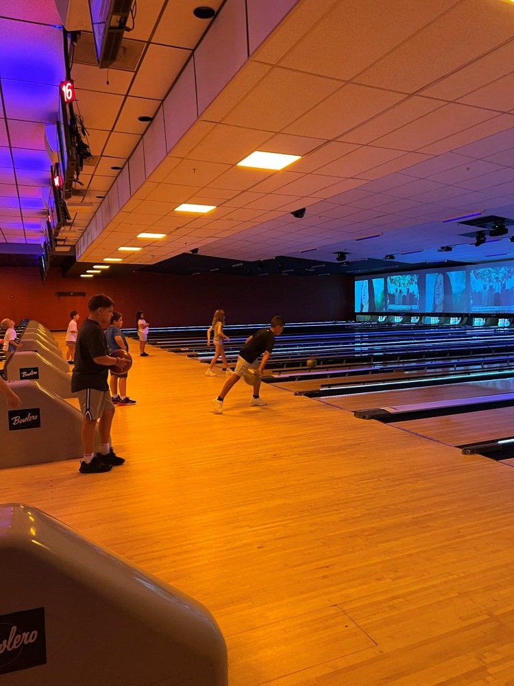 Campers stand along the approach area of a brightly lit bowling alley, each taking turns rolling balls down the lanes on a South Cape Community Center camp field trip